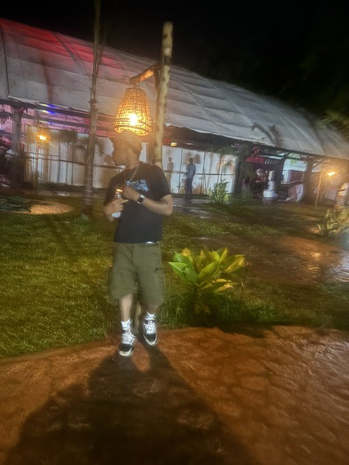 A person in a black t-shirt and green shorts stands on a wet path at night, holding a drink under a hanging wicker lamp near a tented outdoor venue.