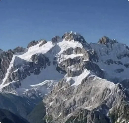 The user wants a caption for the image.
The image shows a mountain range with snow-covered peaks and rocky faces under a blue sky.
The prompt includes a specific note: "如果你是右脑型的，你看到会是一只鲸鱼；如果你是左脑型的，你看到的会是一隻熊猫" (If you are right-brained, you will see a whale; if you are left-brained, you will see a panda).
I need to describe the image concisely (1-2 sentences) and mention that it relates to the text provided, which implies a visual illusion or pareidolia (seeing shapes in clouds/mountains).

Looking at the image:
- It's a mountain range.
- There are snow patches.
- The shapes are jagged and rocky.
- The prompt text suggests seeing a whale or a panda. This is a classic "Rorschach" or pareid