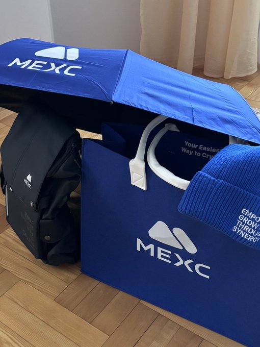 MEXC-branded merchandise including a blue umbrella, tote bag ("Your Easiest Way to Crypto"), backpack, beanie, and shirt displayed on a wooden floor, received as a prize from a contest by @MEXC_Poland and @CryptoPodroze.