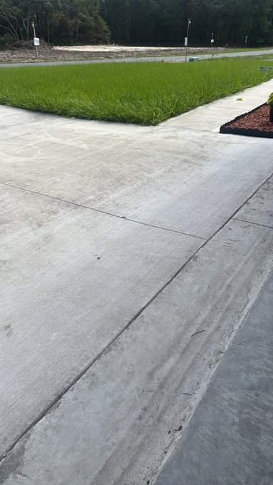 A freshly poured concrete driveway or path stretches alongside a patch of green grass, with construction debris visible in the background.