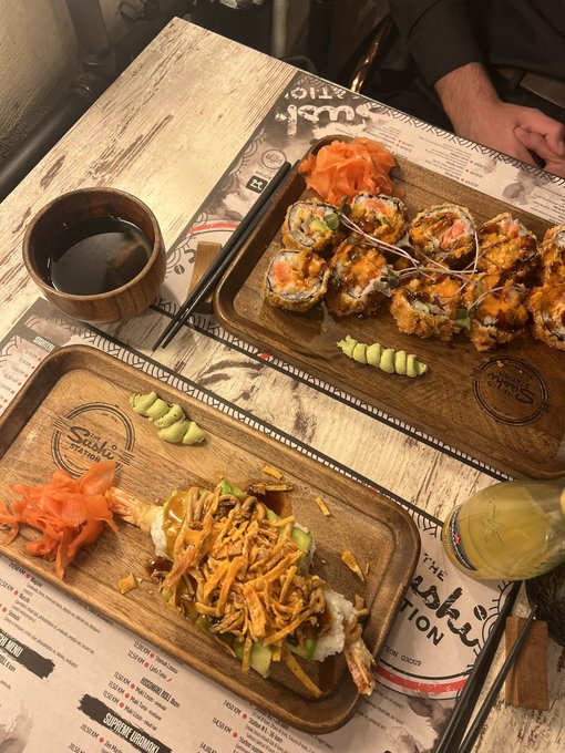 A wooden tray holds sushi rolls topped with fried onions alongside pickled ginger, wasabi, a bowl of soy sauce, and chopsticks on a restaurant table.
