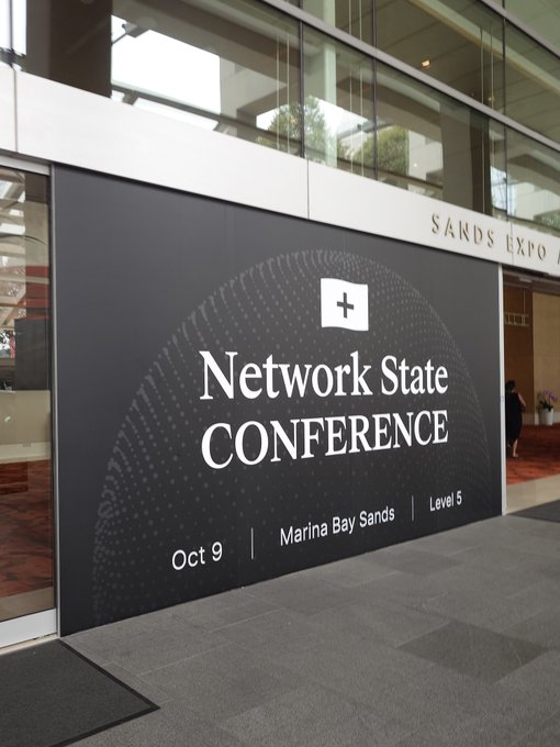 A black event sign at Sands Expo reads 'Network State CONFERENCE' with a white plus logo, advertising the event for October 9 at Marina Bay Sands, Level 5.