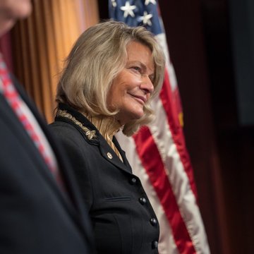 Senator Cynthia Lummis, with blonde hair and wearing a dark blazer, is shown smiling in front of an American flag during what appears to be an official government event, reflecting her determined stance in the post about pressing forward with the CLARITY Act despite the frustrating delay.