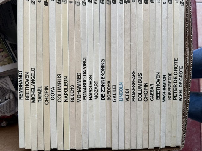 A collection of book spines in a cardboard box, with names of famous artists printed on them including REMBRANDT, BEETHOVEN, MICHELANGELO, RAFAEL, CHOPIN, GOYA, COLUMBUS, NAPOLEON, RUBENS, MOHAMMED, LEONARDO DA VINCI, NAPOLEON, MOZART, DE ZONNEKONING, BOEDDHA, GALILEI, LINCOLN, VERDI, SHAKESPEARE, COLUMBUS, CHOPIN, CAESAR, BEETHOVEN, WASHINGTON, ROBESPIERRE, PETER DE GROTE, and KAREL DE GROTE.