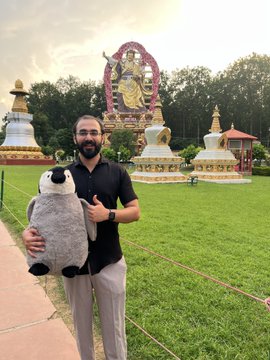 A man with glasses and a beard holding a large panda plush toy, standing on a path with a golden Buddhist statue and white stupas visible in the background, appearing to be at a temple site.