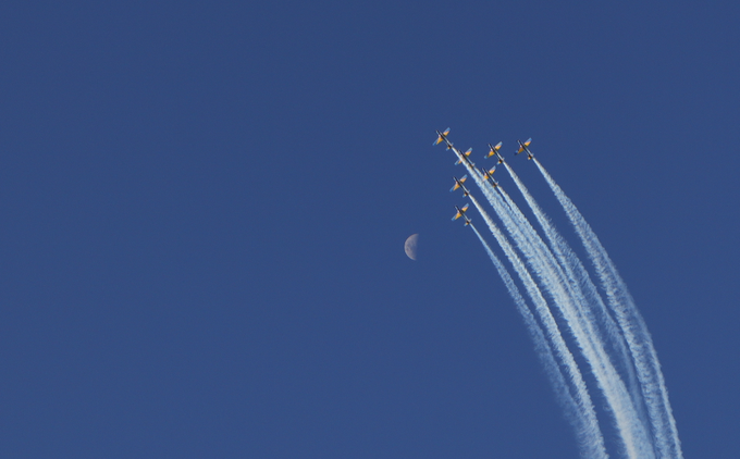 Against a deep blue sky, a formation of jets flies diagonally, leaving parallel white contrails behind them as they pass near a small crescent moon.