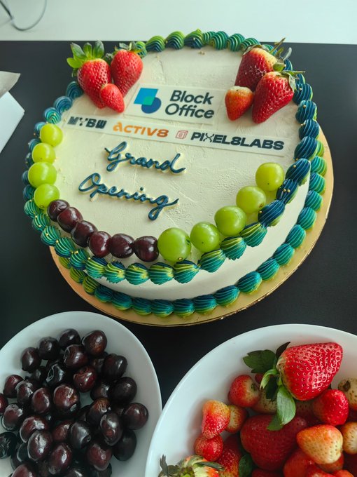 A celebratory "Grand Opening" cake decorated with strawberries, grapes, and green frosting borders features the Block Office logo alongside branding for "M:BI," "ACTIV8," and "PIXEL8LABS," commemorating the joint office launch event held on 17th August 2026.