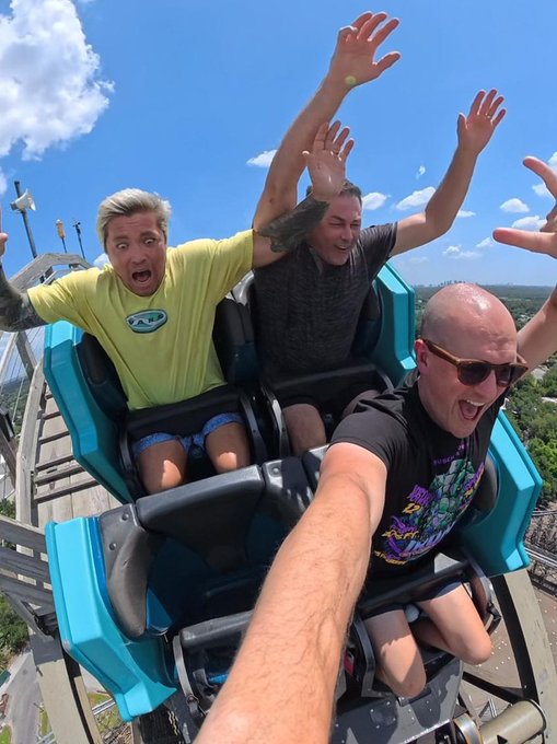 Three friends throw their hands in the air while riding a rollercoaster, with one wearing a yellow shirt featuring an "AEV" logo. The image captures the thrill and freedom of enjoying life's experiences while building wealth through Bitcoin stacking.