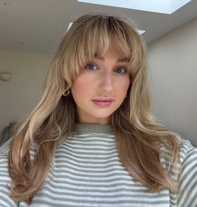 A young woman with long blonde hair, curtain bangs, and a green and white striped sweater looks softly at the camera. Her serene, natural look pairs perfectly with the casual "gm" (good morning) greeting, giving off a cozy, ready-to-start-the-day vibe.