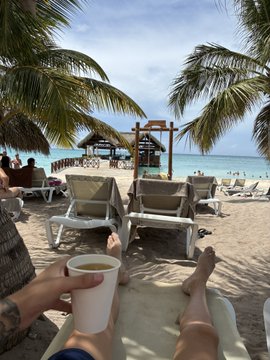 A person's feet resting on a beach lounge chair holding a white cup, with palm trees, ocean, and a cruise ship visible in the background.