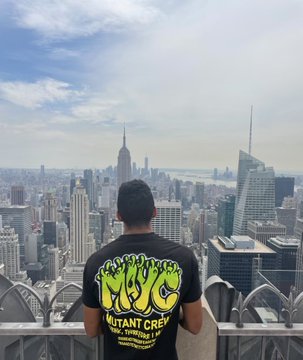 A man stands on an observation deck with his back to the camera, wearing a black t-shirt with green 'MUTANT CREW' graffiti-style graphics, looking out over the New York City skyline including the Empire State Building.