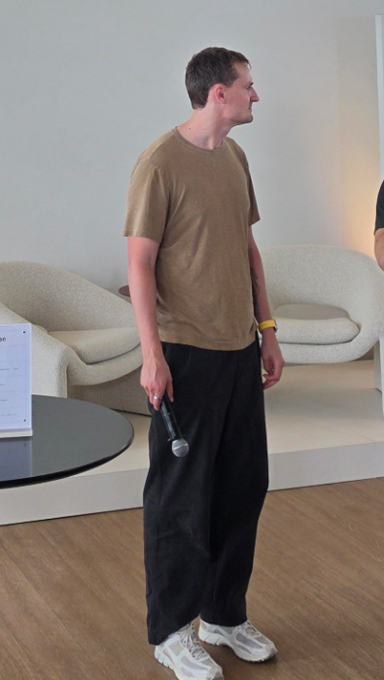 A tall man stands indoors on a wooden floor wearing an oversized tan t-shirt and baggy black pants with white sneakers, holding a microphone in a room with curved beige chairs.