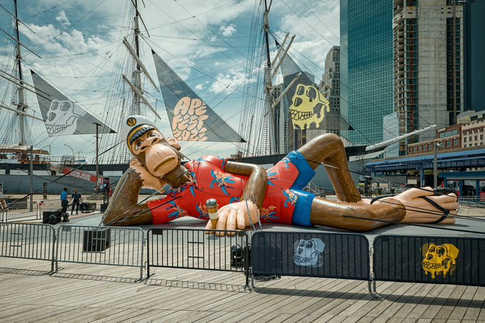 A large inflatable or sculpture of a pirate character lounging on a wooden boardwalk near a docked sailing ship, with city buildings in the background.