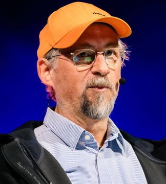 A man with glasses, a gray goatee, and an orange cap (featuring what appears to be a Bitcoin-related logo) wears a headset microphone, likely appearing at a Bitcoin-focused event or conference. The image relates to the post about H100's Bitcoin acquisition, possibly depicting a figure connected to the Bitcoin ecosystem, though no specific name is visible in the image to confirm an exact identity link to Adam Back or H100.