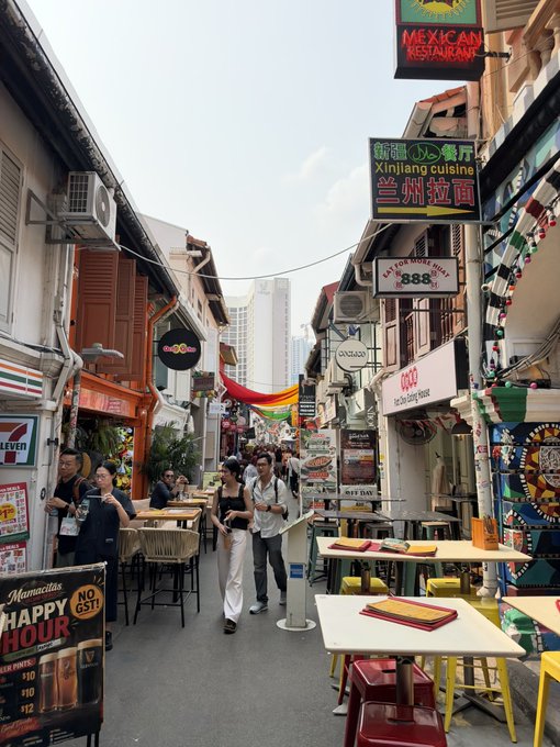 A vibrant street scene in Singapore's Haji Lane area, featuring colorful shophouse signage including 'Xinjiang cuisine', 'MEXICAN RESTAURANT', and 'Maracuya', along with a 'NO GST! Happy Hour' sign and pedestrians enjoying the pedestrian street.