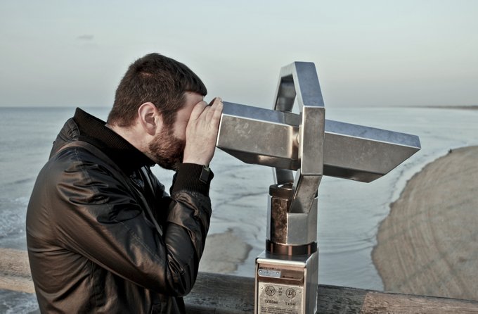 A man with a beard looking through a coin-operated viewing telescope mounted on a railing, with a beach and ocean visible in the background.