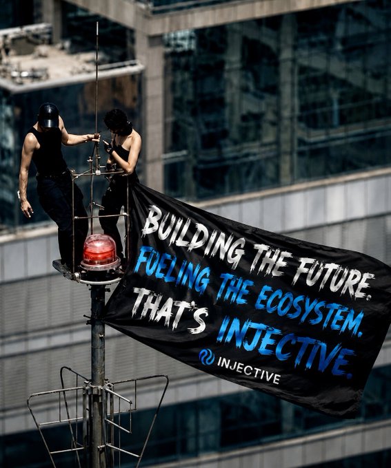 Two activists on a rooftop antenna platform unfurling a black banner reading 'BUILDING THE FUTURE. FUELING THE ECOSYSTEM. THAT'S INJECTIVE.' with the Injective logo.