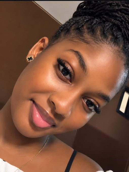 A close-up selfie of a smiling woman with braided hair, wearing a black flower-shaped stud earring.