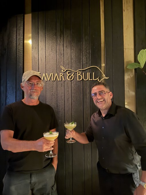 Two men smile and toast with cocktails at Caviar & Bull, with the restaurant's illuminated sign displayed on the dark wood-paneled wall behind them.