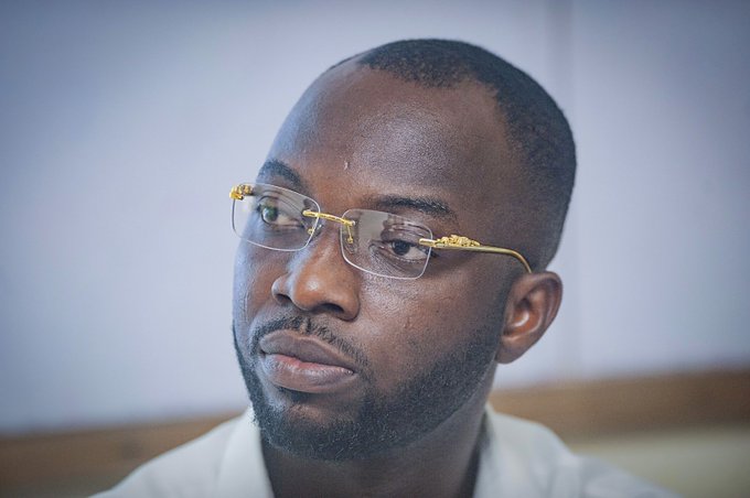 A close-up portrait of a man with a short beard wearing gold-rimmed glasses, looking off to the side.