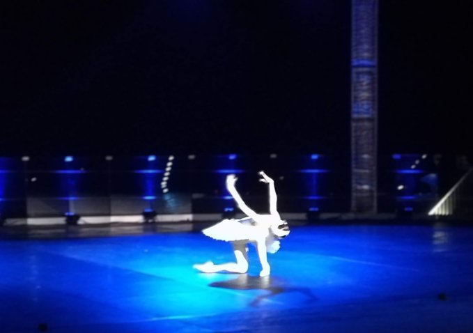 A blurry nighttime image of a ballerina in a white tutu performing a handstand pose on a blue-lit stage, with a tall illuminated tower visible in the background.
