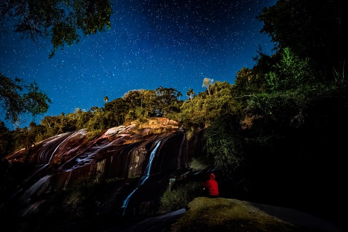 Under a star-filled night sky, a person in a red hoodie sits beside a cascading waterfall, illuminating the rocky landscape with a warm glow. The scene captures the natural beauty and stillness of the moment, reflecting the artwork's blend of hand-animated art and original photography featured in the Still, Not Still NFT collection.