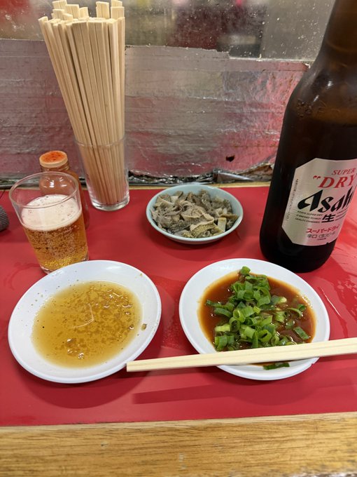 A photo of a meal at a Japanese restaurant, featuring a glass of Asahi Super Dry beer, a glass of water, bamboo chopsticks, and small dishes of garlic oil and scallions, alongside a plate of what appears to be liver sashimi.