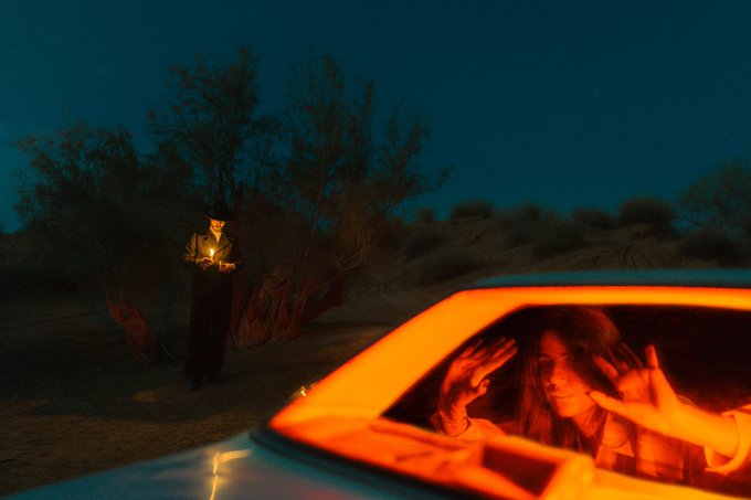 The image shows a car with its headlights on, driving down a road at night. In the background, there are trees that cast long shadows onto the road. A person can be seen standing near one of these trees, holding an object or a lantern. The photo is titled "GN A Whisper to Delirium" and has a caption that reads "0.2 Ξ".