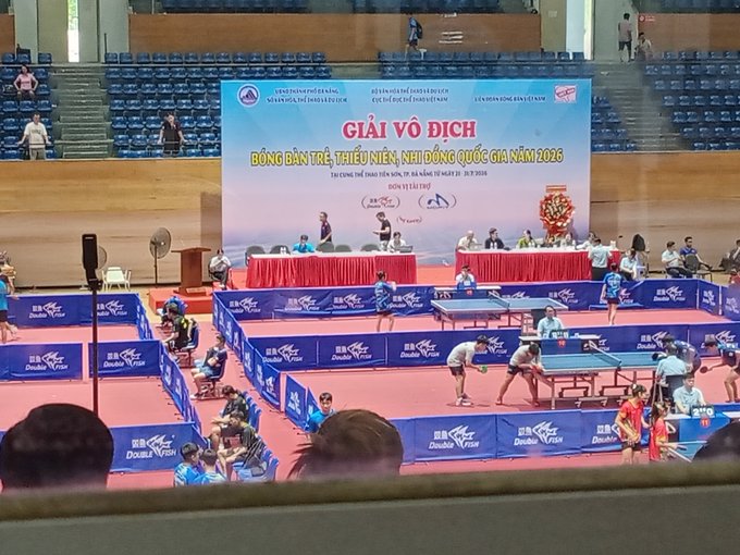 A table tennis tournament is underway with the banner 'GIẢI VÔ ĐỊCH BÓNG BÀN TRẺ, THIẾU NIÊN, NHI ĐỒNG QUỐC GIA NĂM 2026' displayed above the courts.