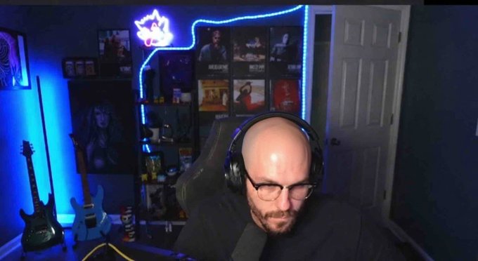 A bald streamer with glasses and headphones sits in a gaming room featuring blue LED lighting, a neon sign, two guitars, and shelves displaying various items and posters, appearing focused during a stream. The image accompanies a post about tipping $500 on stream and running it back, suggesting the streamer is engaged in a live broadcast where viewers can receive tips.