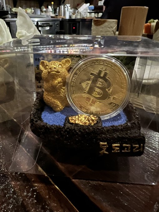 A souvenir display case containing a small golden figurine and a gold-colored Bitcoin commemorative coin on a black base with Korean text reading "롯데코리" (Lotte Co.).