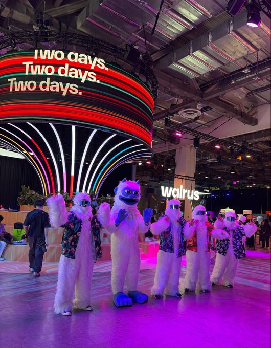 A trade show or convention floor featuring people dressed in white furry yeti costumes wearing sunglasses and colorful Hawaiian shirts, performing or dancing in front of a large curved LED display showing 'Two days. Two days. Two days.' with a 'walrus' sign visible in the background.