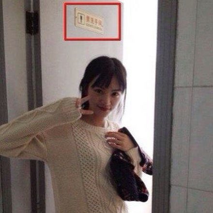 A woman wearing a cream-colored knit sweater poses with a peace sign in front of a doorway, with a framed sign behind her containing partially visible red text.