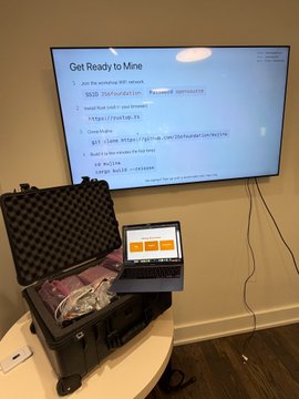 A wall-mounted TV displays a "Get Ready to Mine" presentation slide with the URL "http://mujina.org" and Linux download instructions, while an open hard case below reveals a custom development hashboard with cables, placed in front of an open laptop showing a terminal interface—representing the open-source Bitaxe mining hardware setup for the Mujina project mentioned in the post.