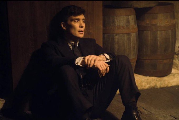 A man in a dark suit sits on the floor leaning against a wall in a dimly lit room with wooden barrels behind him, appearing to be a scene from a film or television show.