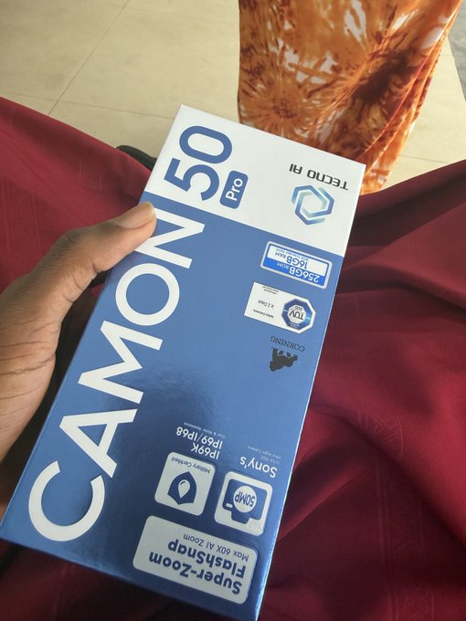 A hand holds a blue product box for a "TECNO AI" "CAMON 50 Pro" smartphone featuring "Super-Zoom 50MP", "Sony IPrv9b", and "256GB+16GB RAM" text against a red fabric background.