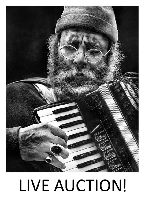 A black-and-white promotional image for a 'LIVE AUCTION!' featuring a bearded man in a beanie and glasses playing an accordion, wearing distinctive rings.