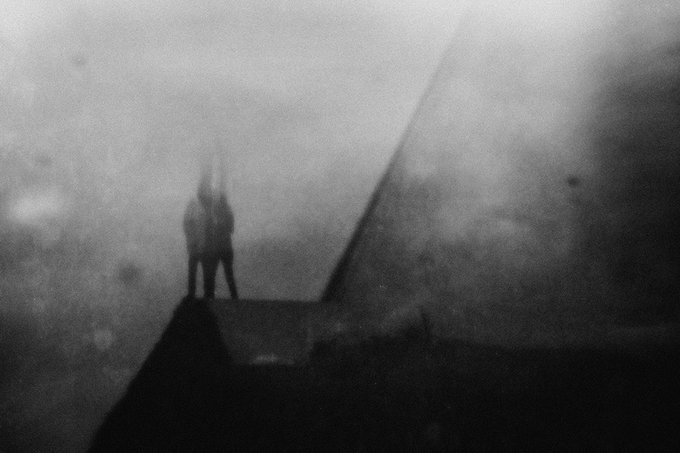 A grainy, dark black-and-white image showing a silhouette of a figure with raised arms standing on a structure, possibly the bow of a ship, against a hazy, obscured background.