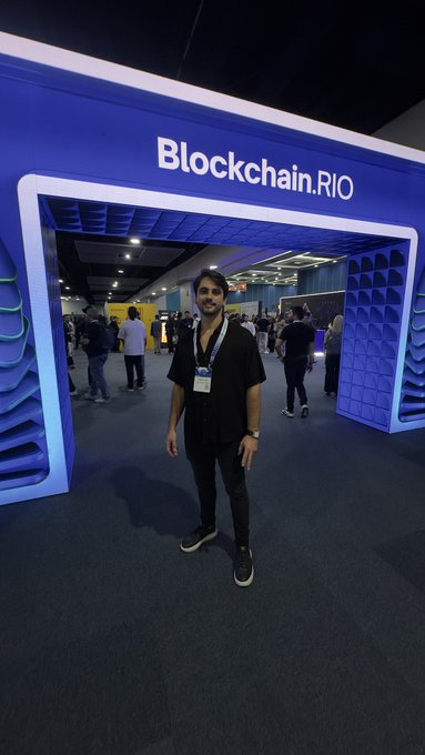 A young man wearing a lanyard standing at the entrance of the 'Blockchain.RIO' conference venue.