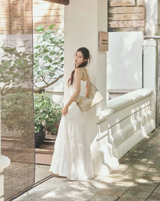 Photo of a woman in a long white dress holding a white handbag, standing on a sunlit patio with bamboo blinds and plants.