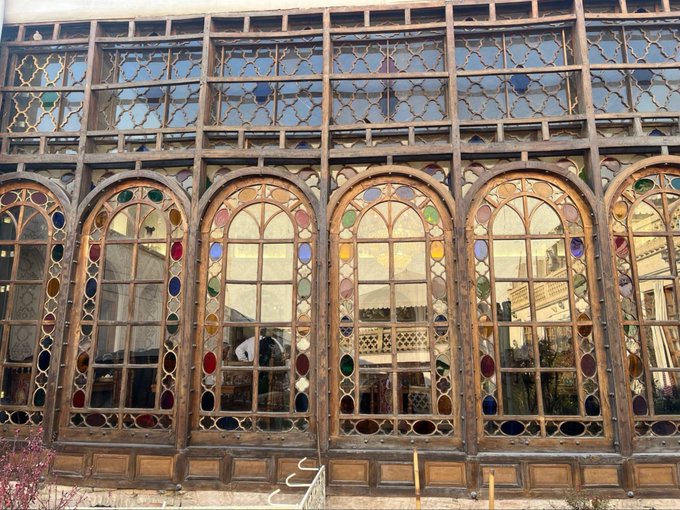 An ornate building facade features large arched windows with intricate wooden frames and colorful stained glass details reflecting the surrounding architecture.