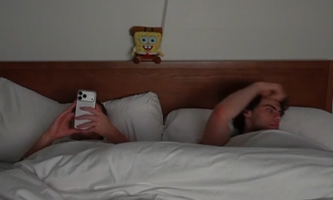 Two people in bed under covers, one looking at a phone screen, with a SpongeBob plush toy on the headboard between them.