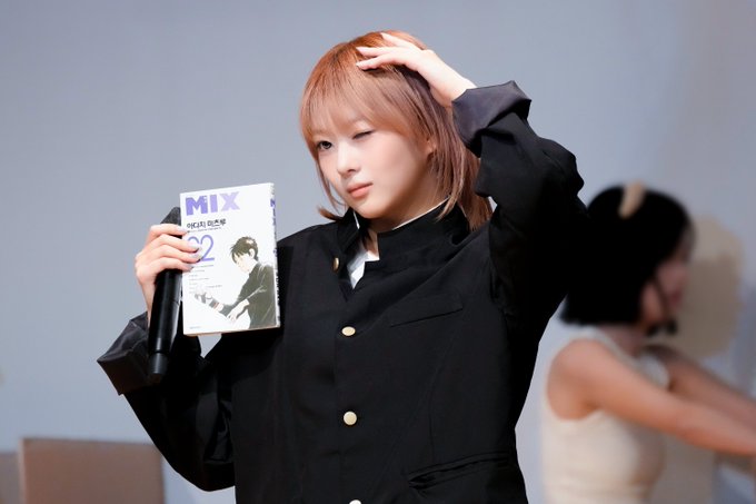A young woman with light brown hair wearing a black jacket with gold buttons, holding up a copy of "MIX" magazine (issue 22) and a microphone while touching her head, with another person blurred in the background.