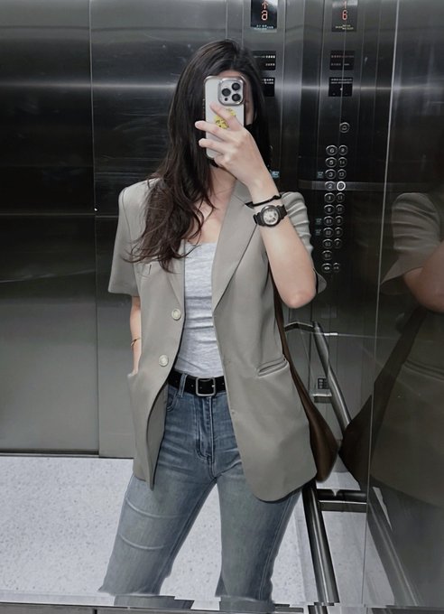 A woman takes a mirror selfie in an elevator, wearing a gray blazer over a white tee and jeans, while the caption humorously laments losing both money and energy. The polished elevator setting contrasts with the self-deprecating "work hard, stay broke" sentiment, emphasizing her exhausted yet stylish composure.