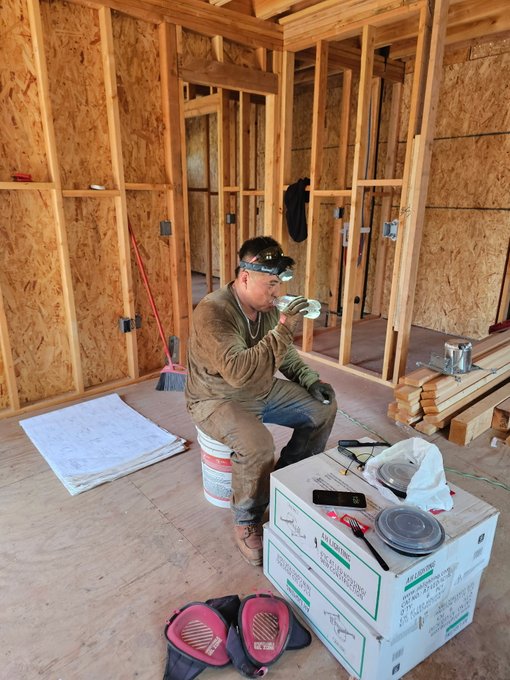 A construction worker wearing a headlamp takes a break to drink from a cup while sitting on a bucket inside a wood-framed building under construction.