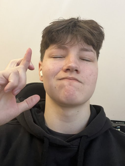 A young man with dark hair wearing a white earbud and a black hoodie closes his eyes and raises his hand in a peace sign against a plain white wall.