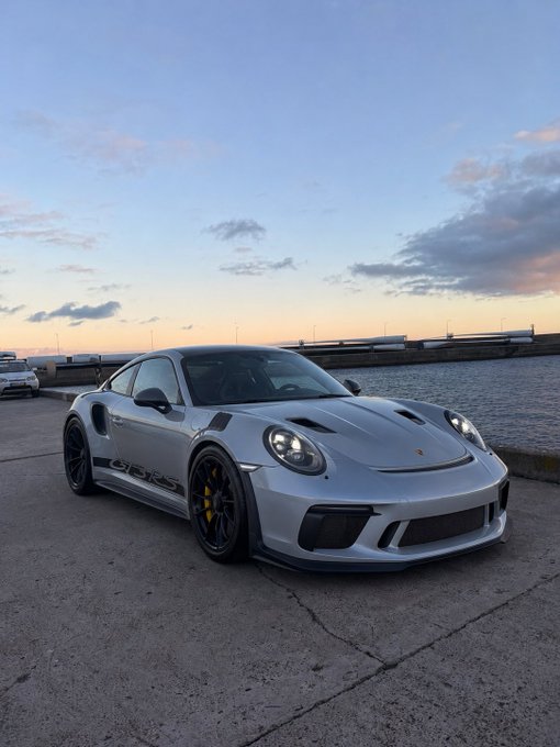 A sleek silver Porsche 911 GT3 RS is parked by the water at sunset, prominently displaying "GT3 RS" badging on its side, fitting a post referencing a $SOL crypto wallet address.