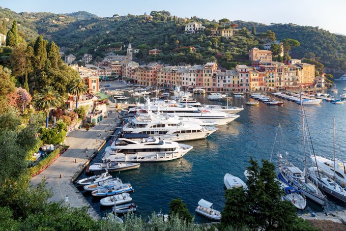 A scenic aerial view of Portofino, Italy, showing colorful buildings lining a harbor filled with yachts and boats, surrounded by lush green hills.
