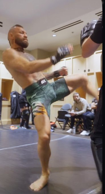 A muscular shirtless man wearing camouflage shorts and MMA gloves executes a high kick during training, embodying the athletic movement quality referenced in the post about Conor McGregor's movement decline due to "decoding."