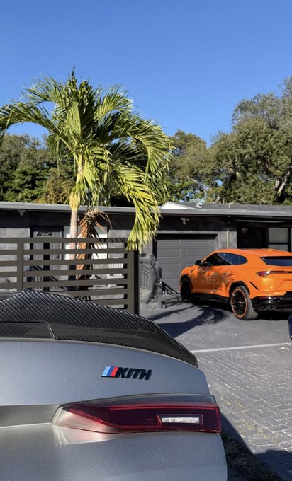 A photograph taken from behind a grey car with a 'KITH' badge, looking towards an orange Lamborghini Urus parked in a driveway next to a palm tree.